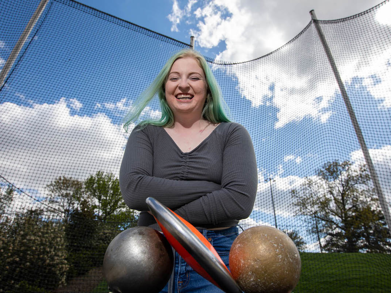 Picture description: picture is of a smiling young white woman with long green hair wearing a long-sleeved gray shirt with arms crossed. She is standing outside with track and field equipment under a blue sky with puffy white clouds.
