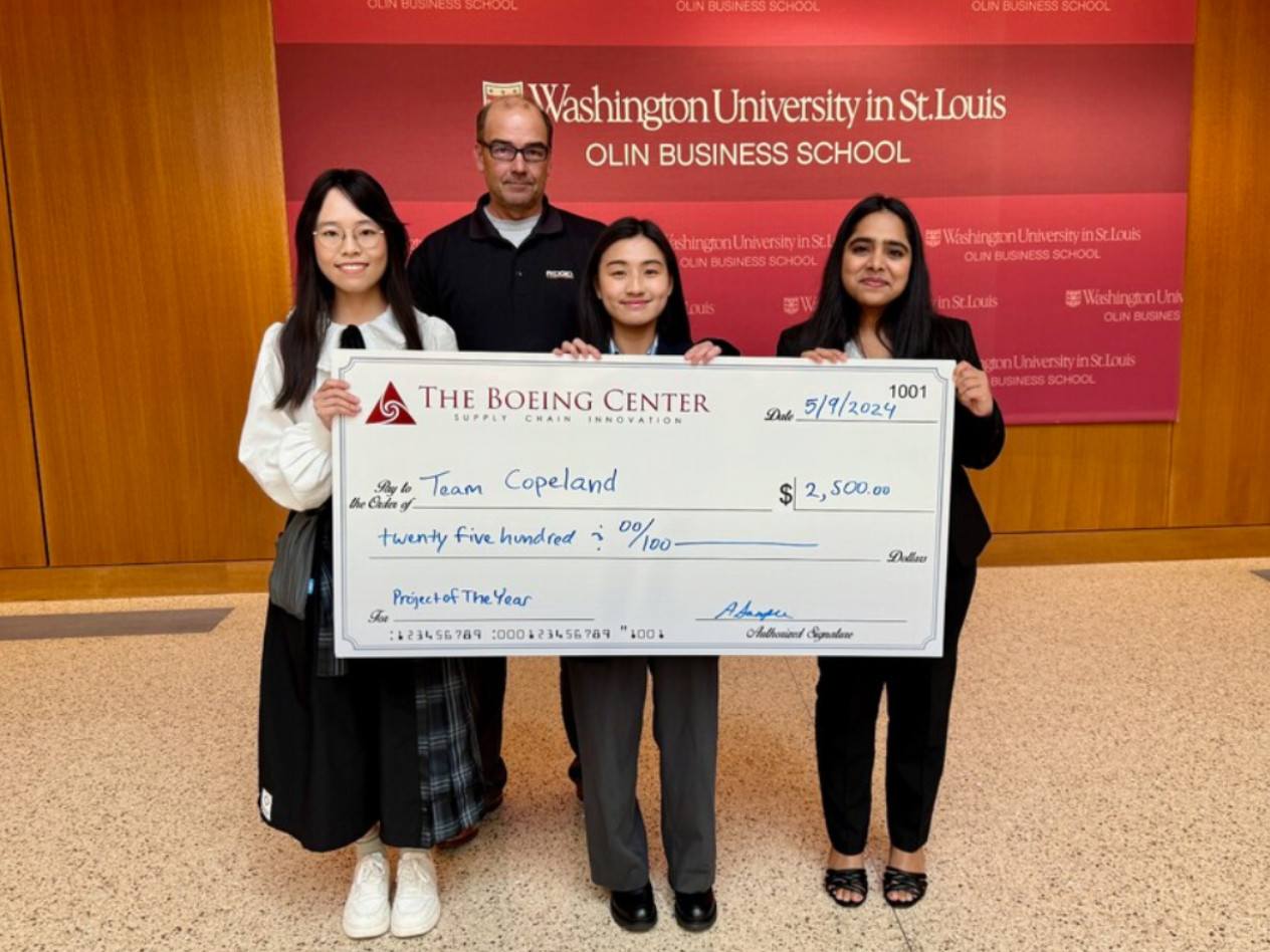 Three students and a client stand holding an oversize check for $2,500