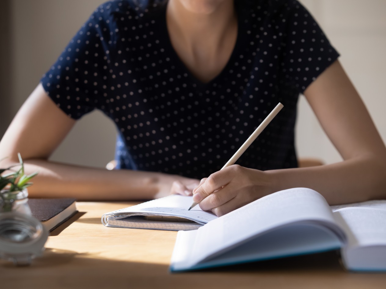 Cut off shot of young woman at desk writing on a pad with a book in front of her.