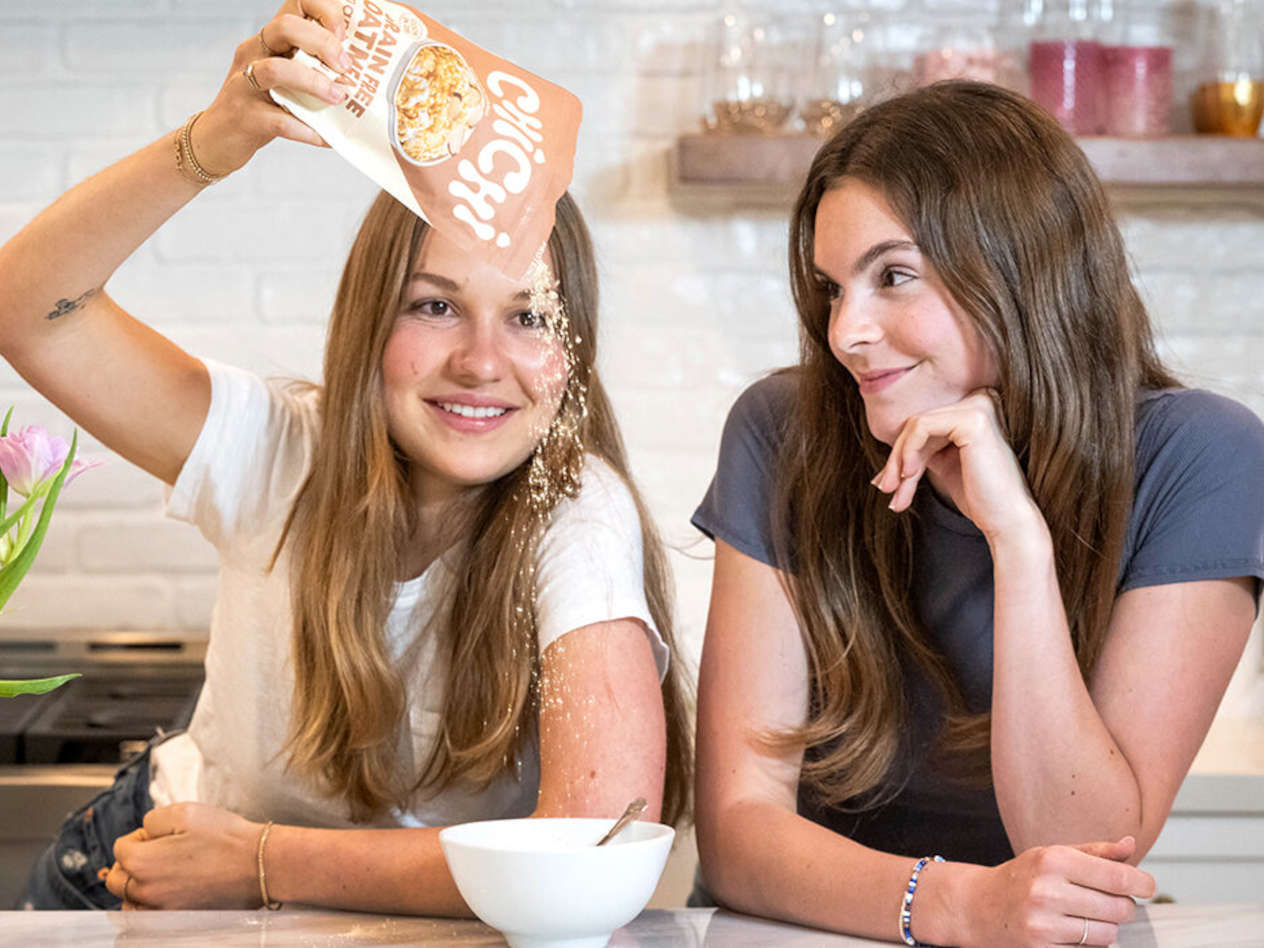 Two young women stand at a counter, one pours a cereal into a bowl.