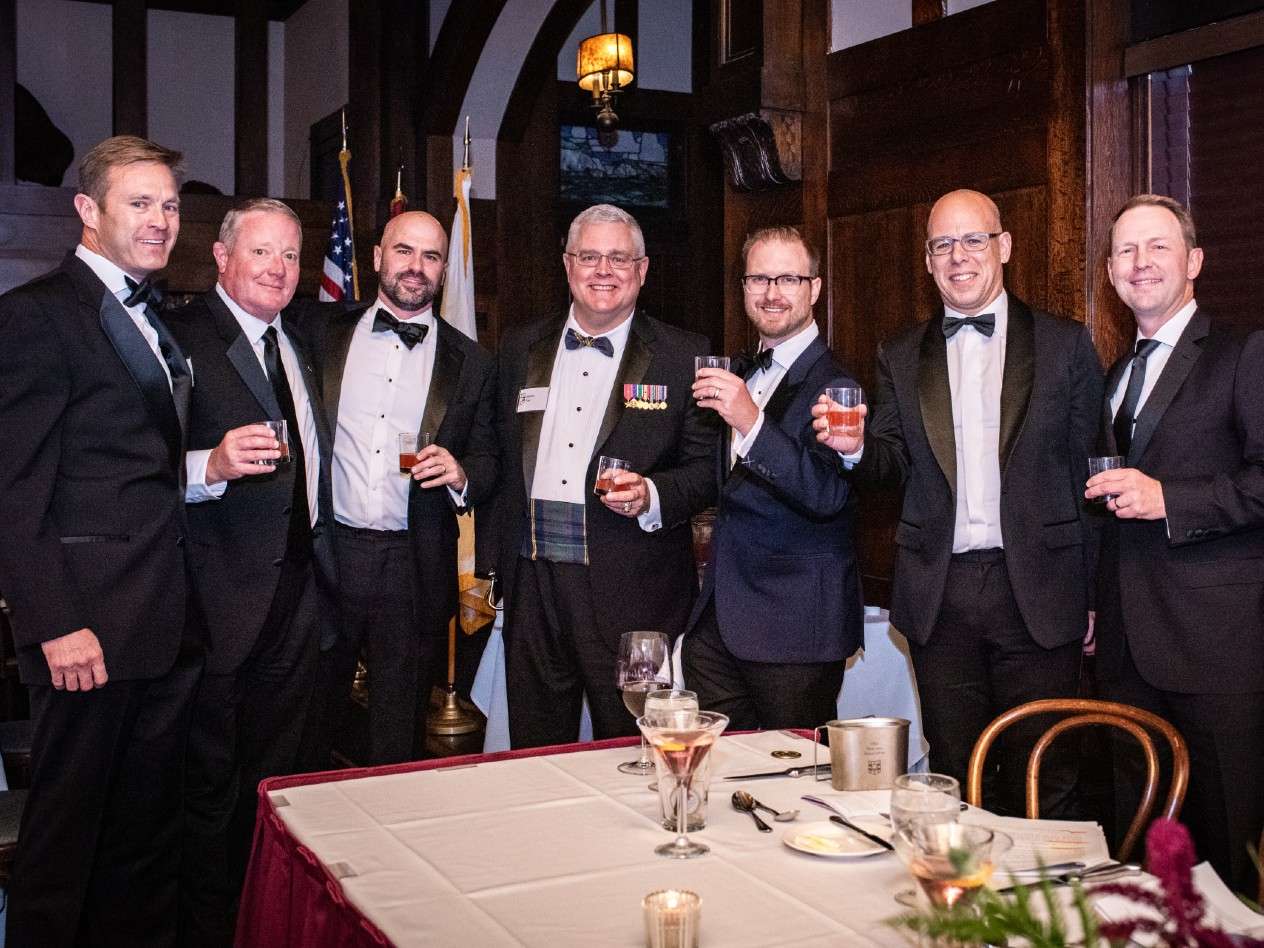 A group of men stand at a formal dinner. one is wearing several medals on his jacket.