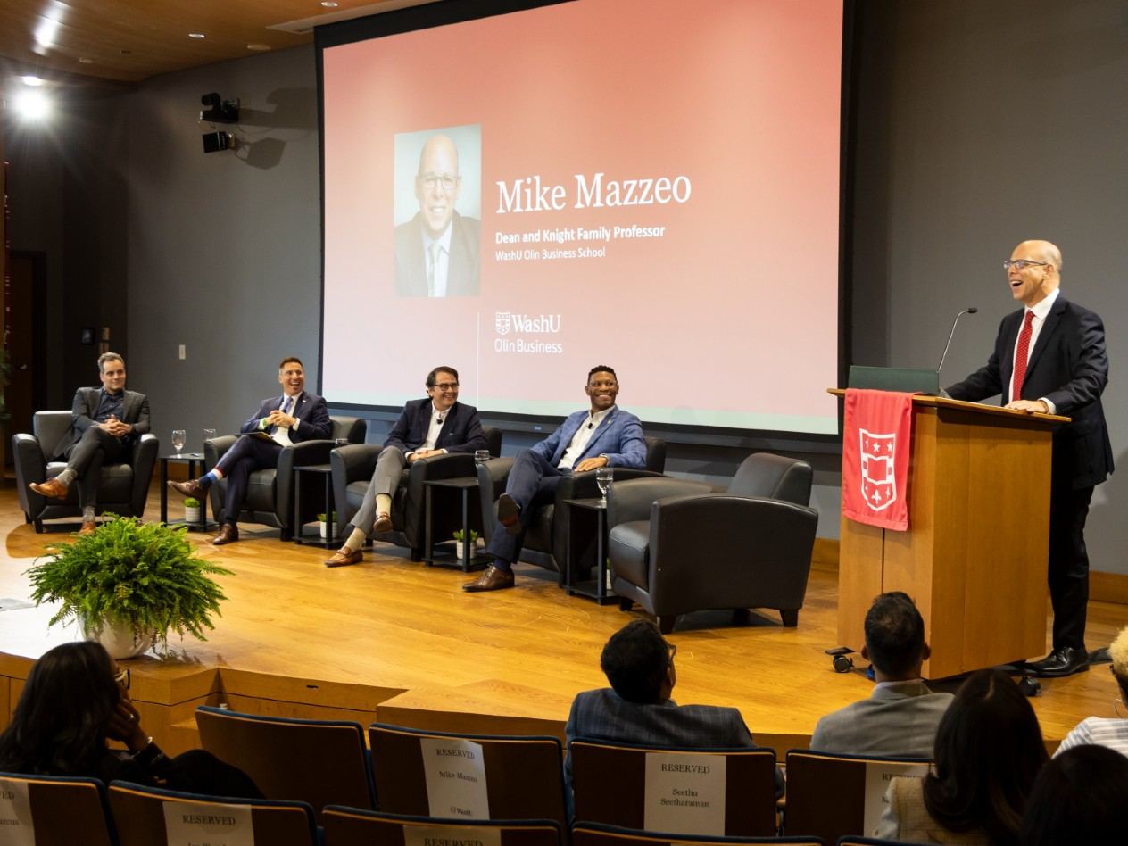 A man stands at a podium, addressing a crowd while four other men sit in chairs on a stage.