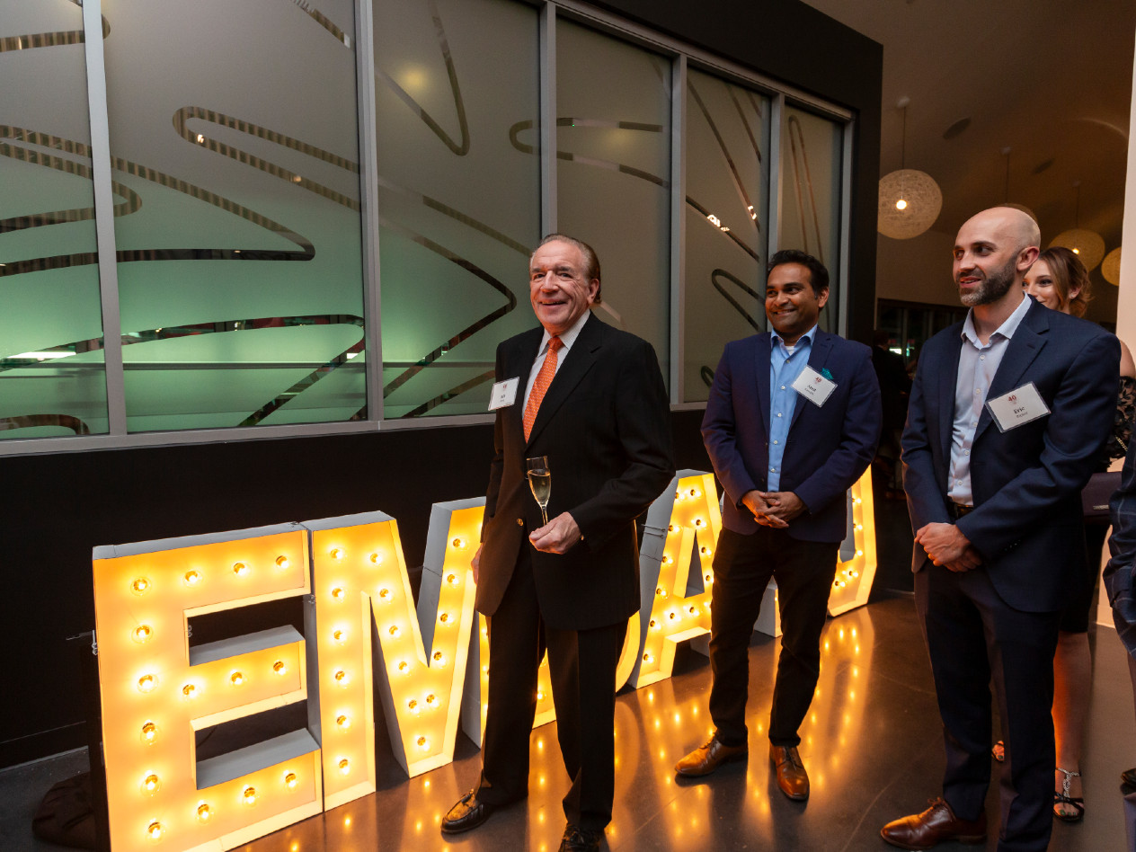 Three men stand in front of a lighted EMBA sign.