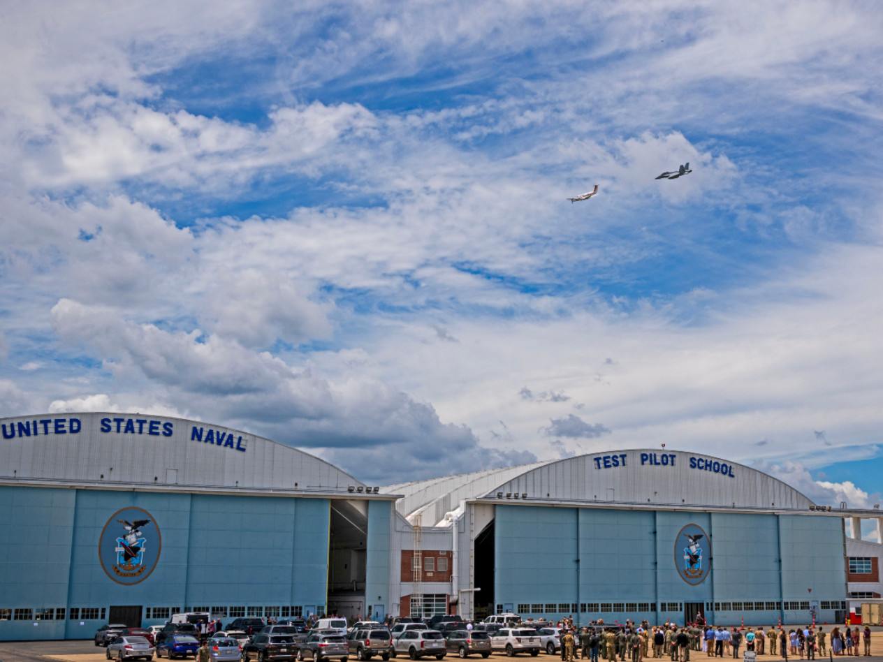 Planes flying above a hangar bearing the words 'United States Naval Test Pilot School'